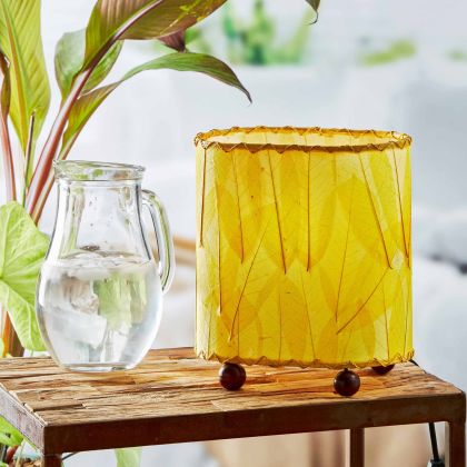 In a softly lit reading nook, crafted by the artisans of Eangee Home Design, the Mini Guyabano Table Lamp in yellow stands gracefully, featuring fossilized cocoa leaf shade, guyabano leaf shade, wrought iron frame, adding warmth and character to the space.