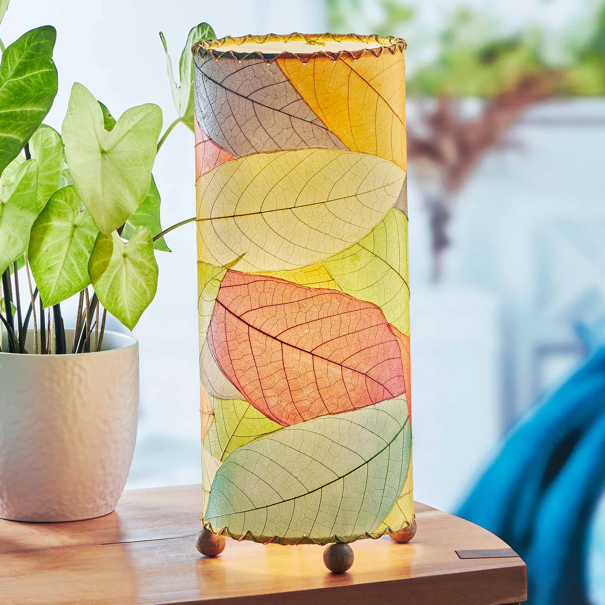 In a modern interior with earth tones, crafted by the artisans of Eangee Home Design, the Cocoa Leaf Cylinder Table Lamp in multi color stands gracefully, featuring fossilized cocoa leaf shade, wrought iron frame, handcrafted, adding warmth and character to the space.