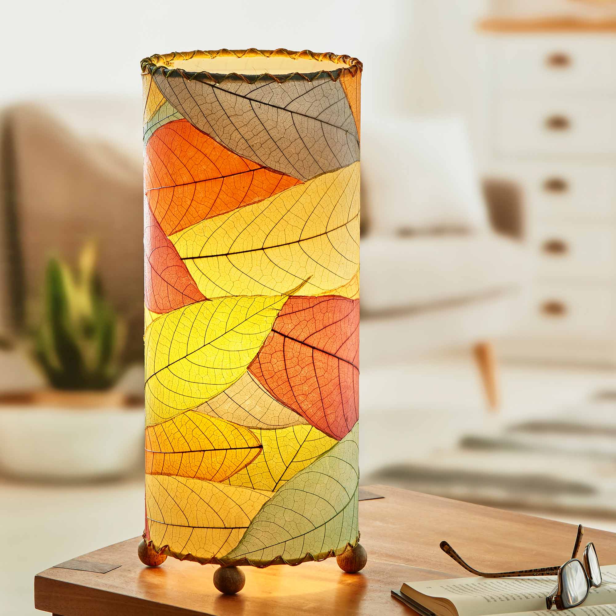 In a softly lit reading nook, crafted by the artisans of Eangee Home Design, the Cocoa Leaf Cylinder Table Lamp in multi color stands gracefully, featuring fossilized cocoa leaf shade, wrought iron frame, handcrafted, adding warmth and character to the space.