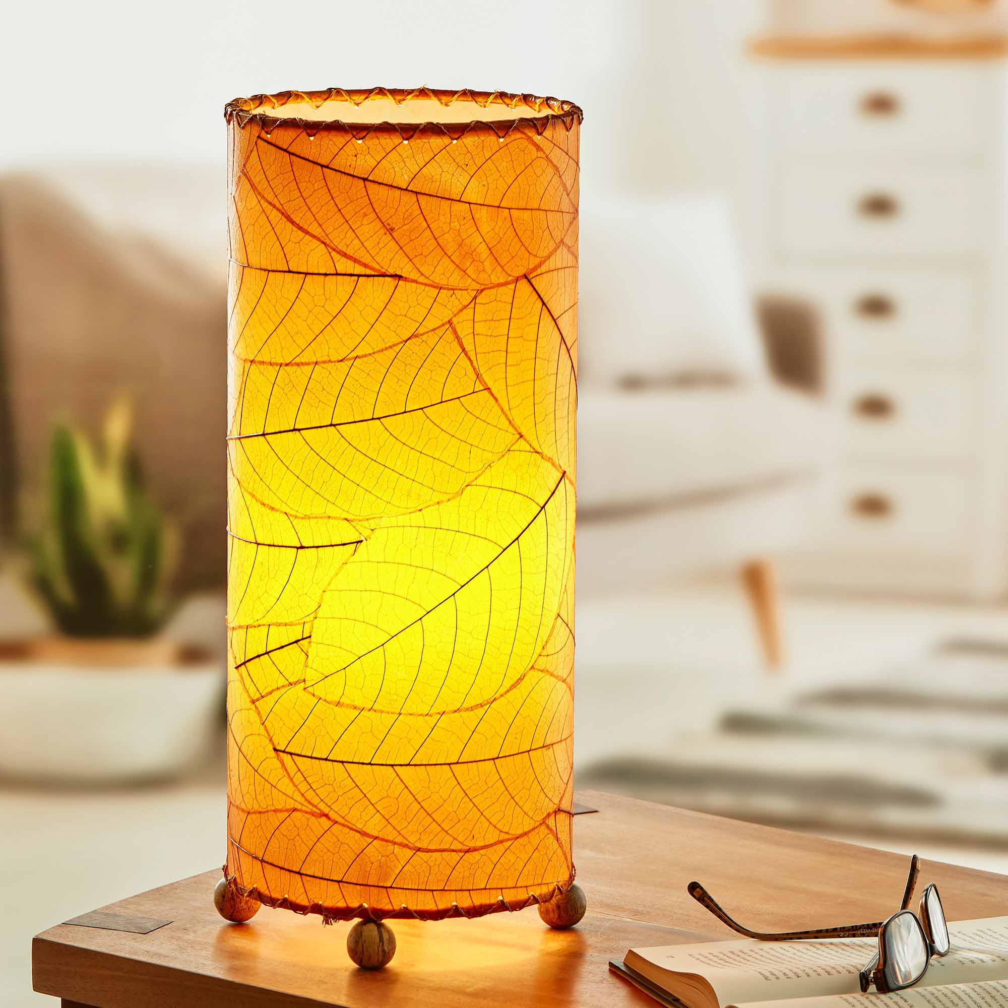 In a modern interior with earth tones, crafted by the artisans of Eangee Home Design, the Cocoa Leaf Cylinder Table Lamp in orange stands gracefully, featuring fossilized cocoa leaf shade, wrought iron frame, handcrafted, adding warmth and character to the space.