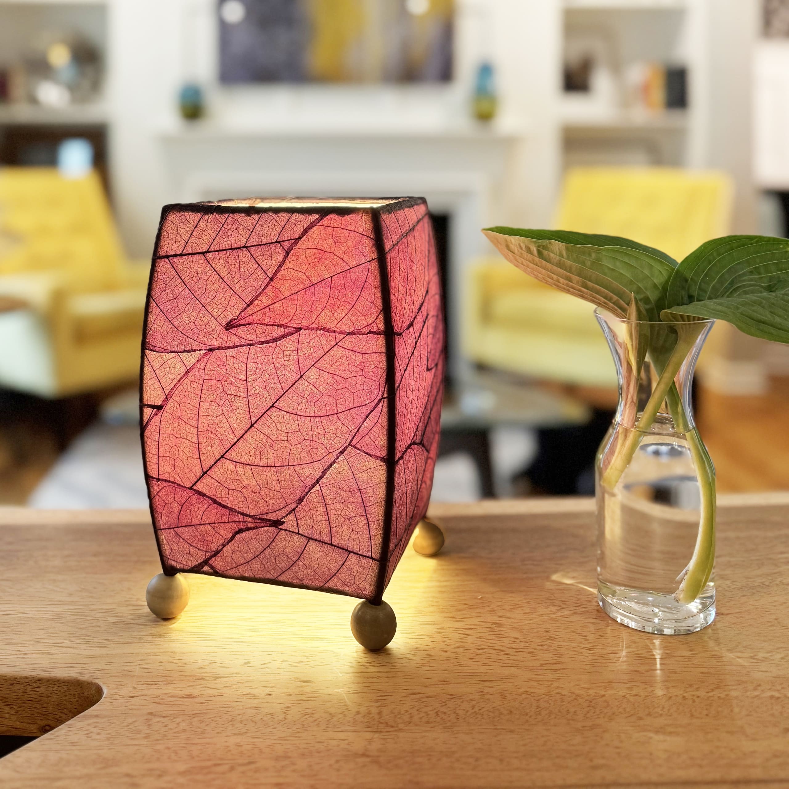 In a coastal inspired living room, crafted by the artisans of Eangee Home Design, the Mini Square Cocoa Leaf Table Lamp in purple stands gracefully, featuring fossilized cocoa leaf shade, wrought iron frame, fair trade, eco friendly, sustainably sourced, handcrafted, adding warmth and character to the space.