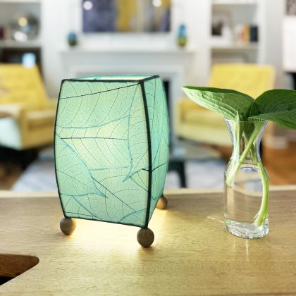 On a bedside table, the Mini Square Cocoa Leaf Table crafted by the artisans of Eangee Home Design Lamp in sea blue finish by Eangee Home Design glows softly, its fossilized cocoa leaf panels, wrought iron frame, and hand placed natural leaves bringing it's the warm, inviting glow that draws you in first.