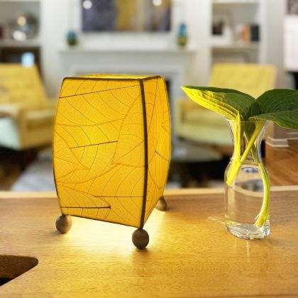 On a floating shelf, the Mini Square Cocoa Leaf Table crafted by the artisans of Eangee Home Design Lamp in yellow finish by Eangee Home Design glows softly, its fossilized cocoa leaf panels, wrought iron frame, and hand placed natural leaves bringing it's the warm, inviting glow that draws you in first.