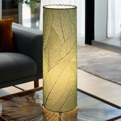In a softly lit reading nook, crafted by the artisans of Eangee Home Design, the 24 Inch Cocoa Leaf Cylinder Table Lamp in green stands gracefully, featuring fossilized cocoa leaf shade, fair trade, eco friendly, sustainably sourced, handcrafted, adding warmth and character to the space.