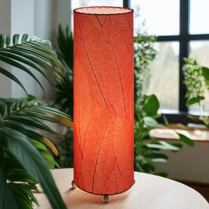 In a softly lit reading nook, crafted by the artisans of Eangee Home Design, the 24 Inch Cocoa Leaf Cylinder Table Lamp in red stands gracefully, featuring fossilized cocoa leaf shade, fair trade, eco friendly, sustainably sourced, handcrafted, adding warmth and character to the space.