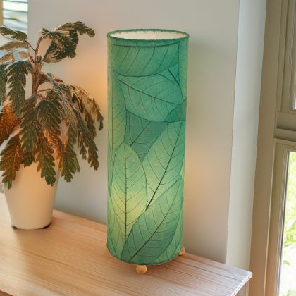 In a quiet bedside corner, crafted by the artisans of Eangee Home Design, the 24 Inch Cocoa Leaf Cylinder Table Lamp in sea blue stands gracefully, featuring fossilized cocoa leaf shade, fair trade, eco friendly, sustainably sourced, handcrafted, adding warmth and character to the space.