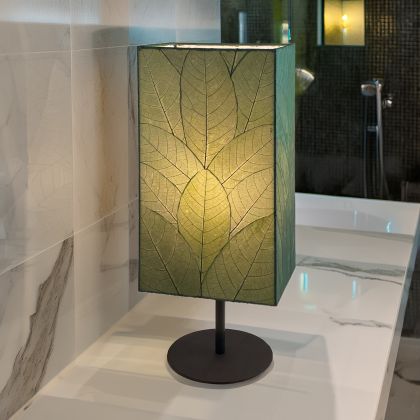 In a softly lit reading nook, crafted by the artisans of Eangee Home Design, the Sequoia Table Lamp in green stands gracefully, featuring fossilized cocoa leaf shade, fair trade, sustainably sourced, adding warmth and character to the space.