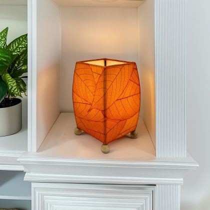 On a wooden console, the Mini Square Cocoa Leaf Table crafted by the artisans of Eangee Home Design Lamp in orange finish by Eangee Home Design glows softly, its fossilized cocoa leaf panels, wrought iron frame, and hand placed natural leaves bringing it's the warm, inviting glow that draws you in first.