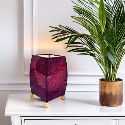 On a floating shelf, the Mini Square Cocoa Leaf Table crafted by the artisans of Eangee Home Design Lamp in purple finish by Eangee Home Design glows softly, its fossilized cocoa leaf panels, wrought iron frame, and hand placed natural leaves bringing it's the warm, inviting glow that draws you in first.