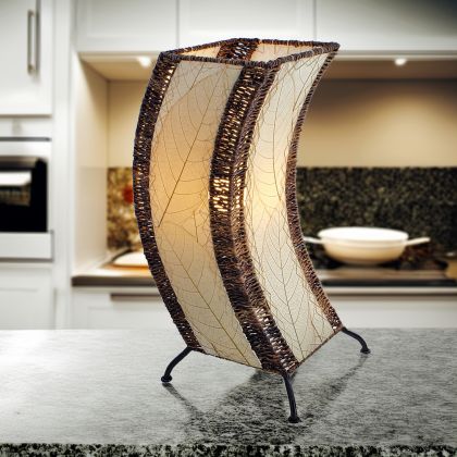 In a softly lit reading nook, crafted by the artisans of Eangee Home Design, the C Shape Table Lamp in natural stands gracefully, featuring fossilized cocoa leaf shade, abaca rope accents, fair trade, handcrafted, adding warmth and character to the space.