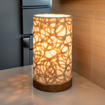 In a quiet bedside corner, crafted by the artisans of Eangee Home Design, the Mini Paper Cylinder Table Lamp Swirl in natural stands gracefully, featuring reclaimed mango wood base, recycled paper shade, sustainably sourced, recycled materials, adding warmth and character to the space.