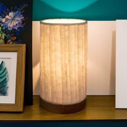 In a softly lit reading nook, crafted by the artisans of Eangee Home Design, the Mini Paper Cylinder Table Lamp Lines in natural stands gracefully, featuring reclaimed mango wood base, recycled paper shade, sustainably sourced, recycled materials, adding warmth and character to the space.