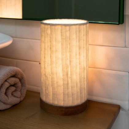 In a softly lit reading nook, crafted by the artisans of Eangee Home Design, the Mini Paper Cylinder Table Lamp Lines in natural stands gracefully, featuring reclaimed mango wood base, recycled paper shade, sustainably sourced, recycled materials, adding warmth and character to the space.