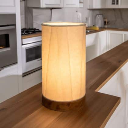 In a softly lit reading nook, crafted by the artisans of Eangee Home Design, the Mini Paper Cylinder Table Lamp Glacier in natural stands gracefully, featuring reclaimed mango wood base, recycled paper shade, sustainably sourced, recycled materials, adding warmth and character to the space.