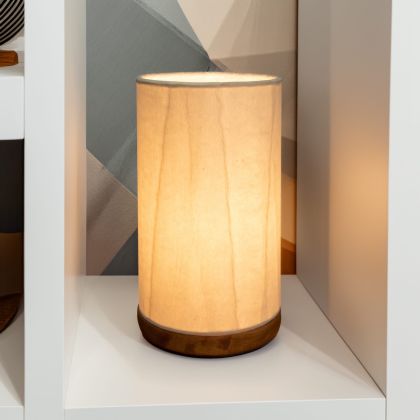 In a softly lit reading nook, crafted by the artisans of Eangee Home Design, the Mini Paper Cylinder Table Lamp Glacier in natural stands gracefully, featuring reclaimed mango wood base, recycled paper shade, sustainably sourced, recycled materials, adding warmth and character to the space.