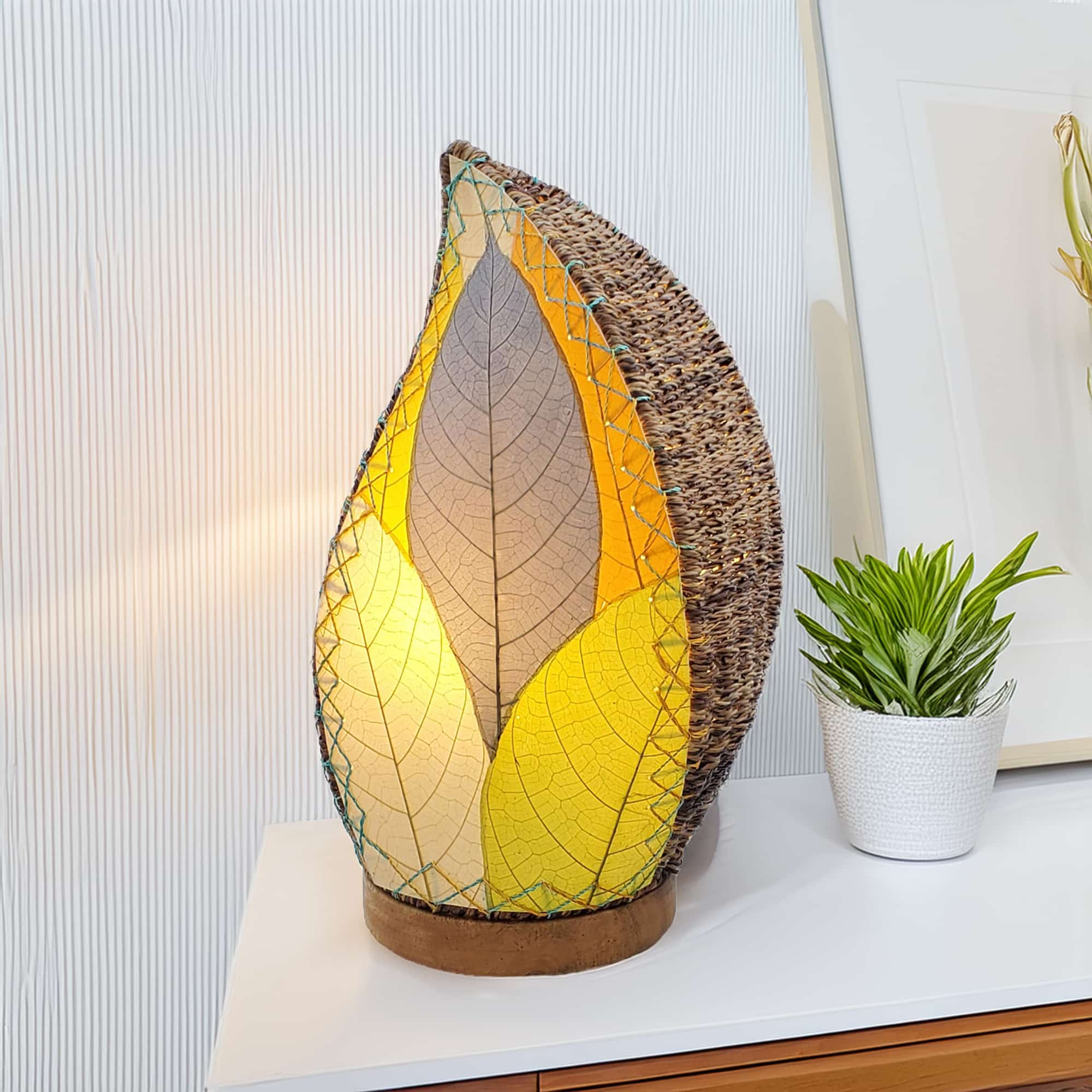 In a quiet bedside corner, crafted by the artisans of Eangee Home Design, the Leaflet Table Lamp in multi color stands gracefully, featuring fossilized cocoa leaf shade, abaca rope accents, reclaimed mango wood base, wrought iron frame, sustainably sourced, adding warmth and character to the space.