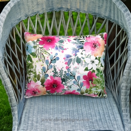 In an outdoor reading nook a multi colored outdoor pillow crafted by the artisans of Magnolia Casual called the Outdoor Pillow Meadow Dance 1 19x24 is displayed featuring 100 percent weather resistant fabric adding comfort and style to the space.