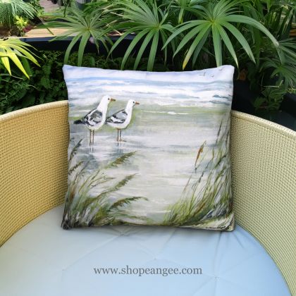 In a sunlit patio lounge a multi colored outdoor pillow crafted by the artisans of Magnolia Casual called the Outdoor Pillow Seagulls on Beach 18x18 is displayed featuring quality outdoor fabric adding comfort and style to the space.
