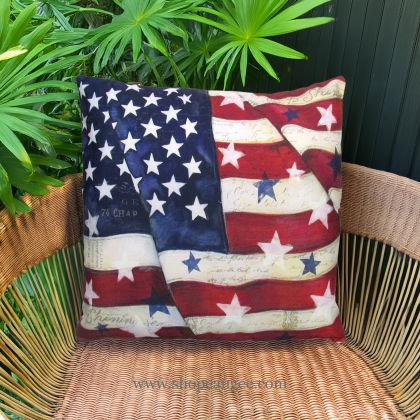 In an outdoor reading nook a multi colored outdoor pillow crafted by the artisans of Magnolia Casual called the Outdoor Pillow American Flag with Stars 18x18 is displayed featuring quality outdoor fabric adding comfort and style to the space.