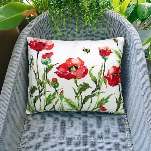 In an outdoor reading nook a multi colored outdoor pillow crafted by the artisans of Magnolia Casual called the Outdoor Pillow Poppies and Bee 19x24 is displayed featuring handwoven textile adding comfort and style to the space featuring with a bee motif.