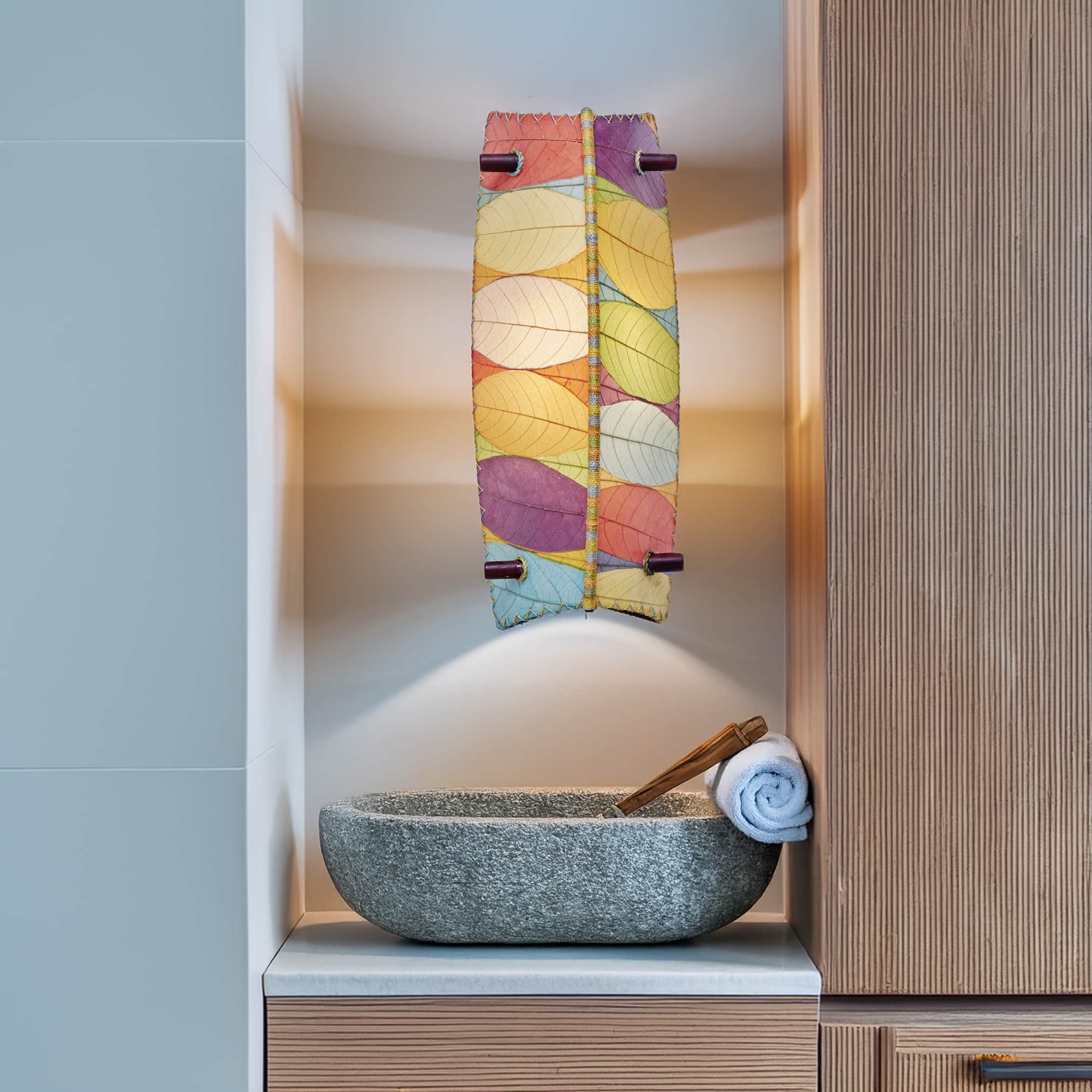 Eangee Home Design Banana Wall Sconce in multicolor with a shade made of real cocoa leaves, casting vibrant light above a stone vessel sink in a modern bathroom niche