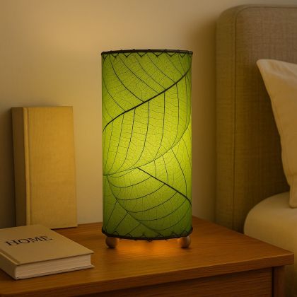 12 Inch Cocoa Leaf Cylinder Table Lamp Green shade, handcrafted from real cocoa leaves . soft ambient light on a bedside table with books, adding a calming green ambiance.