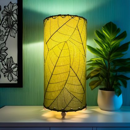 12 Inch Cocoa Leaf Cylinder Table Lamp Green shade, handcrafted from real cocoa leaves . soft ambient light on an accent cabinet against a teal wall beside a leafy plant.