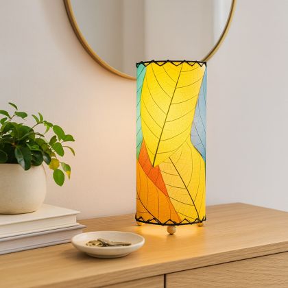 12 Inch Cocoa Leaf Cylinder Table Lamp Multi Color shade, handcrafted from real cocoa leaves . soft ambient light on a glossy white coffee table by tall windows in a modern living room.