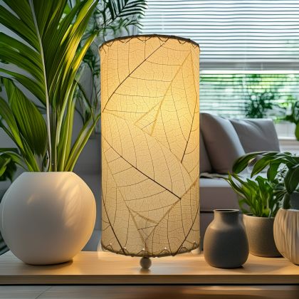 12 Inch Cocoa Leaf Cylinder Table Lamp Natural shade, handcrafted from real cocoa leaves . soft ambient light in a bright living room with houseplants and blinds in the background.