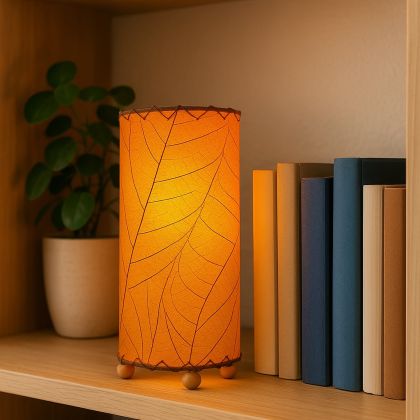 Sustainable orange mini lamp with leaf pattern shade, glowing warmly on a wooden shelf next to books and a small plant.