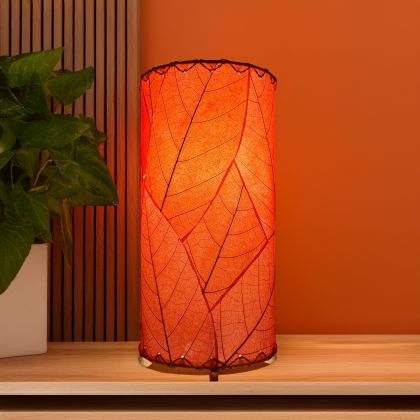 Eangee red mini cylinder lamp with bold cocoa leaf design, casting warm light against an orange wall with a leafy plant beside it.